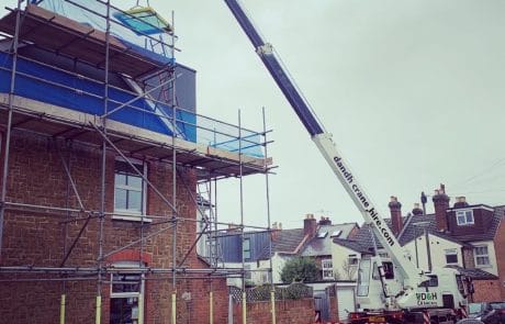 A large D&H Crane Hire crane lifting a heavy glass panel up to a modern loft conversion on a stone house, with the building fully enclosed in scaffolding.